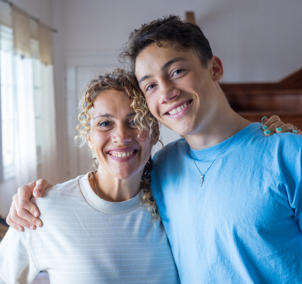 A teenage boy stands with arm around his mother as both smile at the camera.